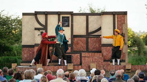 Three actors on a stage outside, dressed in red, blue and yellow costumes from left to right are performing a comical sword fighting scene as the crowd watches on.